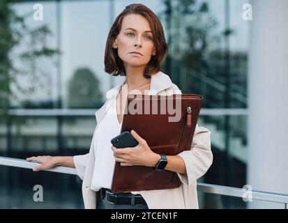 Donna d'affari sicura di mezza età che guarda lontano. Donna in abiti formali che tiene in mano una cartella di pelle mentre si trova all'aperto. Foto Stock