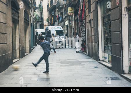 Un ragazzino rimbalza un pallone da calcio su una strada acciottolata nel quartiere Gotico di Barcellona. Vivace e tipica strada di Barcellona Foto Stock