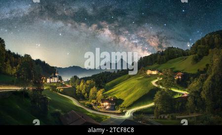 Foto notturna mozzafiato del paesaggio iconico delle Alpi, un idilliaco villaggio con una piccola chiesa e una strada tortuosa sulle colline illuminata da sentieri luminosi Foto Stock