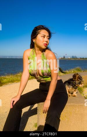 Wind Blown Windy Beautiful Curvy Athletic Asian Woman e Yorkie Dog seduti vicino alla baia di San Francisco Foto Stock