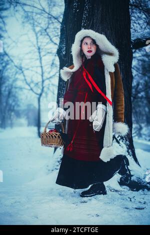 Una graziosa ragazza con un abito rosso folcloristico e un cappotto di pelliccia di pecora con un cesto di mele sta camminando attraverso una misteriosa foresta innevata. Fiaba e slavo f Foto Stock