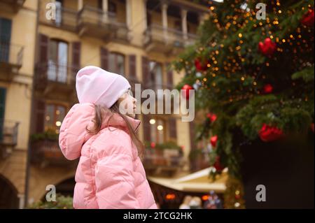 Adorabile bambina vestita con un caldo piumino rosa e un cappello, che fanno un desiderio prezioso, in piedi per strada, ammirando un bellissimo albero di Natale, dicembre Foto Stock
