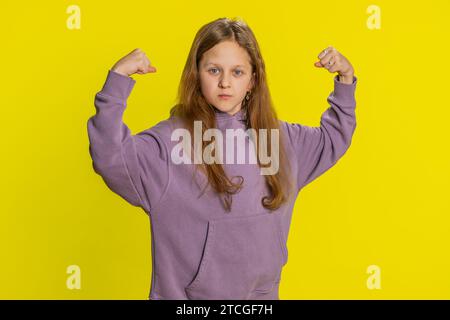 Sono forte e in salute. Giovane ragazza carina che mostra bicipiti e sembra fiduciosa sentendo la forza di lottare per i diritti, l'energia per ottenere il successo vince. Bambino preteen rossa su sfondo giallo Foto Stock