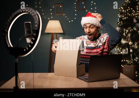 Felice blogger musulmano che indossa il cappello di Babbo Natale disimballando i regali delle confezioni regalo e facendo streaming in diretta. Arabo che usa la fotocamera per smartphone Foto Stock