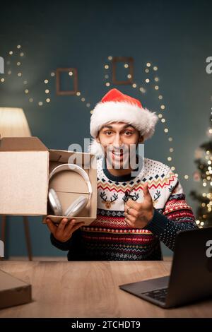 Disimballaggio del video. L'uomo sorridente arabo in un cappello di Babbo Natale a casa mostra ai suoi abbonati nuove cuffie durante una trasmissione in diretta. Concetto pubblicitario influencer Foto Stock