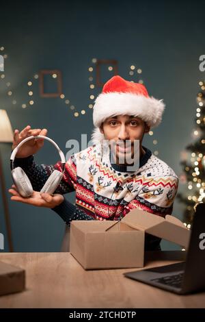 Bel blogger arabo maschio che disimballò un pacchetto con cuffie wireless che registra contenuti video per i social network a casa Foto Stock