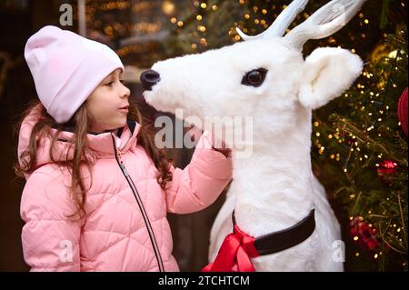 Bella bambina in caldi abiti invernali ammirando un giocattolo di cervi bianchi, in piedi accanto all'albero di Natale, illuminata da ghirlande e luci natalizie, divertimento Foto Stock