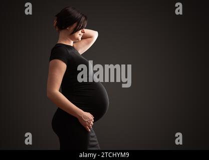 Giovane bella donna incinta con capelli scuri su sfondo scuro Foto Stock