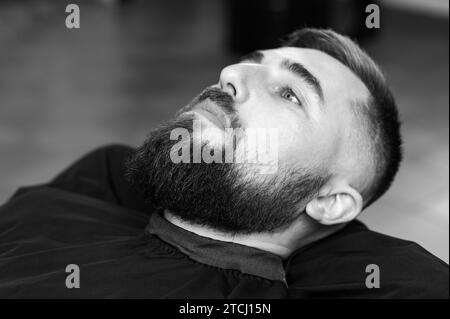 Un giovane con la barba in un mantello nero siede su una sedia in un barbiere. Il cliente sta aspettando un taglio di capelli e la rasatura della barba. Foto Stock