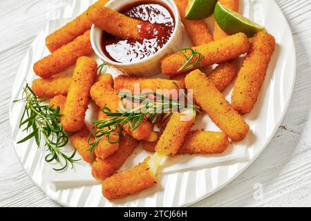 bastoncini di mozzarella impanati fritti serviti con salsa, fette di lime e rosmarino fresco su piatto bianco su tavola di legno bianco con ingredienti, chiudere Foto Stock