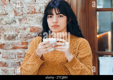 ritratto di una giovane donna latina in piedi accanto a un muro di pietra, è premurosa a bere caffè e a guardare la macchina fotografica e il concetto di caffetteria Foto Stock