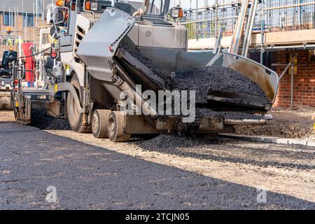 Asfaltatrice riempita con asfalto caldo che posa una nuova superficie stradale sul nuovo sito di sviluppo di abitazioni residenziali e operatore stradale in arancione hi-viz Next Foto Stock