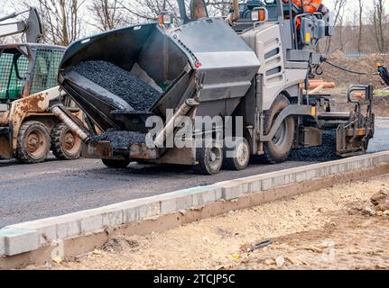 Asfaltatrice riempita con asfalto caldo che posa una nuova superficie stradale sul nuovo sito di sviluppo di abitazioni residenziali e operatore stradale in arancione hi-viz Next Foto Stock