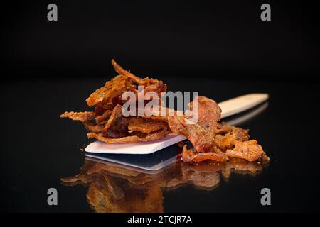 una manciata di carne secca marinata con spezie su un cucchiaio di legno, isolata su uno sfondo nero. Spuntino con birra. Foto Stock