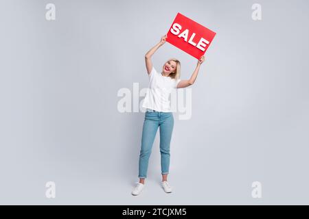 Foto a grandezza naturale di una bella donna di buon umore vestita di t-shirt bianca con un cartello sopra la testa che annuncia la vendita isolata su sfondo di colore bianco Foto Stock