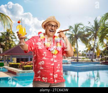 Uomo in piedi vicino a una piscina, fuma un sigaro e tifo con un bicchiere di cocktail Foto Stock