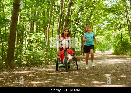Giocosa ragazza che corre con un ragazzo disabile in bicicletta reclinata su un sentiero nella foresta Foto Stock