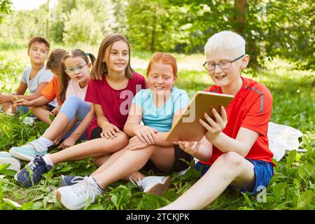 Un ragazzo sorridente che condivide il tablet PC con amici maschi e femmine mentre è seduto al parco Foto Stock