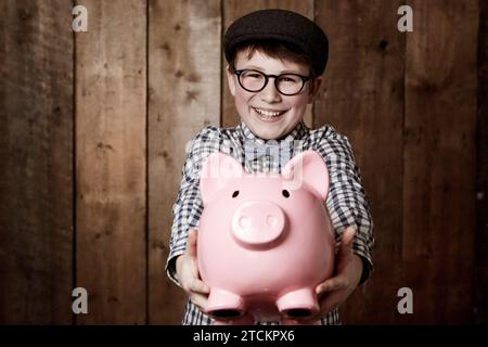 Bambino, salvadanaio o entusiasta di ritratti per sfondo dello studio, risparmi o lezioni di finanza in gioventù. Ragazzo, sorridi e faccia in moneta di piccolo cambiamento Foto Stock