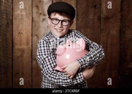 Bambino piccolo, abbraccio e salvadanaio in un ritratto in studio, cassaforte in denaro retrò e risparmi in denaro giovanile. Ragazzo, sorridi e faccia per la banca a moneta Foto Stock