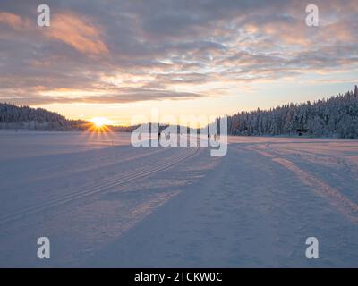 Paesaggio invernale, tramonto su una foresta innevata e un lago ghiacciato in Lapponia. Sciatori e persone che camminano sullo sfondo. Foto Stock