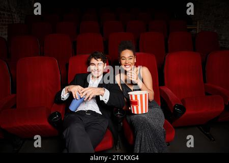 coppia allegra e varia con abiti neri che reggono soda e popcorn e sorridono con gioia Foto Stock