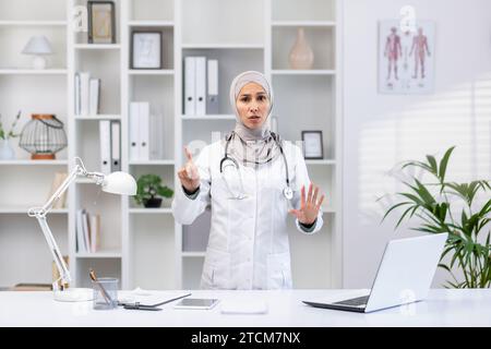 Medico donna allarmata nell'hijab che mostra un segnale di stop con la mano, in piedi in un moderno ufficio privato, guardando la macchina fotografica. Foto Stock