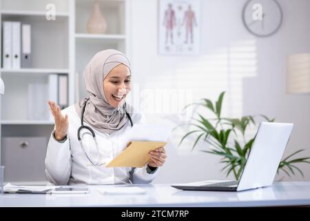 Felice medico femminile in hijab tenendo la lettera in mano, seduto al tavolo in ufficio dell'ospedale, felice di buone notizie. Foto Stock