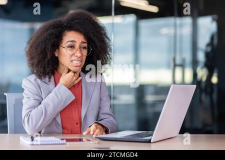 Infelice donna d'affari afroamericana che soffre di mal di gola e tosse, che tiene la mano sul collo, lavora in ufficio. Foto Stock