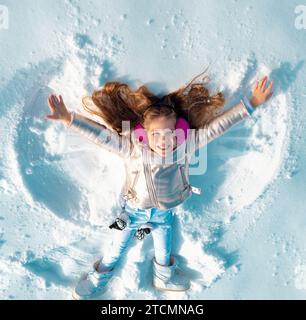 Capretto divertente che fa l'angelo della neve. Bambina che gioca e fa un angelo di neve nella neve. Vista dall'alto. Foto Stock