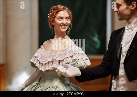 Girate in vita il ritratto di una donna sorridente che indossa un abito da ballo che cammina mano nella mano con un partner gentiluomo nella sala del palazzo, nello spazio fotocopie Foto Stock