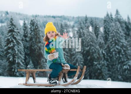 Bambino ragazzo slittando in inverno, giocando con la palla di neve. Bambini in inverno a cavallo sugli scivoli di neve. Inverno foresta di natale con neve caduta e alberi. Foto Stock