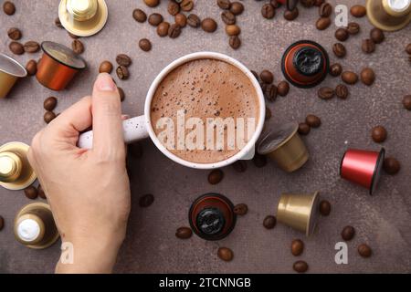Donna che regge una tazza di caffè al tavolo marrone ruvido con capsule e chicchi, vista dall'alto Foto Stock