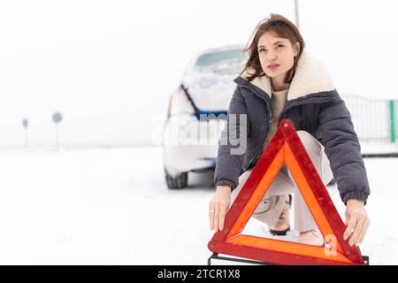 La donna mette un triangolo d'allarme su una strada innevata in inverno, un guasto alle auto in inverno Foto Stock