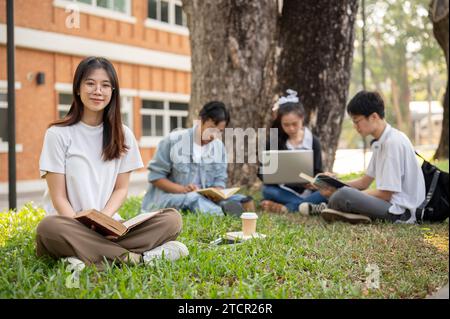 Una giovane studentessa asiatica bella e positiva con un libro è seduta sull'erba in un parco universitario con i suoi amici. Educazione e amicizia Foto Stock