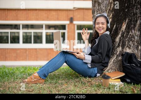 Una giovane studentessa asiatica che indossa delle cuffie sta usando il suo computer portatile sotto l'albero in un parco universitario. immagine vista laterale Foto Stock