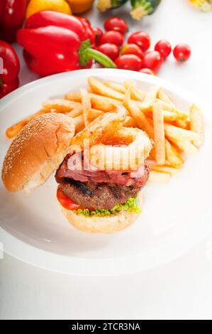 Classico hamburger americano con anelli di cipolla e patatine fritte, con verdure fresche sullo sfondo, CIBO PIÙ DELIZIOSO SU PORTAFOGLIO Foto Stock