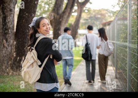 Una graziosa e sorridente studentessa asiatica con lo zaino sta camminando su un sentiero nel suo campus, andando a scuola o tornando a casa dopo le lezioni. Indietro Foto Stock