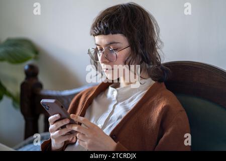 Ragazza hipster pensiva con occhiali in stile vintage che legge le informazioni sullo schermo dello smartphone. Foto Stock
