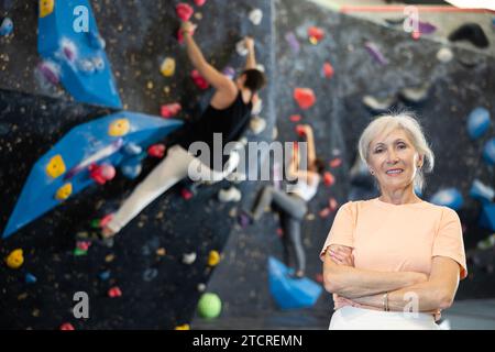 Donna anziana che indossa abbigliamento sportivo sorride alla macchina fotografica mentre si trova in piedi contro l'allenamento artificiale arrampicata parete di roccia nel parco avventura Foto Stock