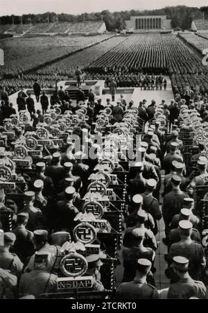 1935 Reichsparteitag (raduno del partito nazista) durante la "Standartenweihe und Totenehrung" (Consacrazione degli standard e commemorazione dei morti), Adolf Hitler è visto fare un discorso davanti a migliaia di truppe e sostenitori. Questo evento, combinando il solenne ricordo dei membri del partito caduti con la consacrazione cerimoniale degli standard militari, evidenzia l'uso del regime di rituali e spettacoli. Il discorso di Hitler, sullo sfondo di una massiccia assemblea, sottolinea la natura orchestrata di questi raduni nel promuovere l'ideologia nazista e rafforzare il ruolo centrale del Führer. Foto Stock