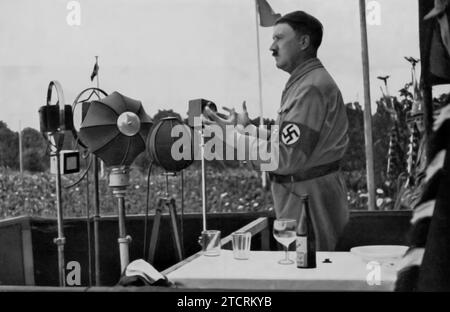 Adolf Hitler è raffigurato rivolgendosi al popolo tedesco, un'immagine che rappresenta i suoi frequenti discorsi pubblici che erano centrali per la sensibilizzazione e la propaganda del regime nazista. Questi discorsi, spesso trasmessi a livello nazionale, furono fondamentali nel trasmettere le politiche e l'ideologia nazista direttamente alla popolazione. Gli indirizzi di Hitler furono progettati per fare appello ai sentimenti nazionali, radunare il sostegno agli obiettivi del regime e rafforzare la sua posizione di leader unificante della Germania. Foto Stock