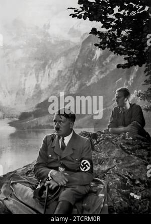 Adolf Hitler, seduto su una roccia a Obersee vicino a Berchtesgaden, con le maestose montagne sullo sfondo. Dietro di lui si trova una forte figura maschile, che probabilmente simboleggia una Germania robusta e protettiva. Questa composizione, ambientata in un paesaggio naturale e scenografico, potrebbe essere intesa a riflettere i temi della forza, della leadership e della connessione dell'ideologia nazista con la patria tedesca. L'ambientazione serena contrasta con l'intensità tipica associata a Hitler, con l'obiettivo di presentare un'immagine più contemplativa e fondata di lui, mentre la figura dietro rafforza il messaggio del potere e della tutela. Foto Stock