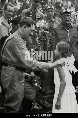 In "Sie sagt ihr Gedichtchen" (recita la sua piccola poesia), questa immagine cattura una giovane ragazza che recita una poesia ad Adolf Hitler durante una delle sue campagne elettorali nel 1932. Questo momento riflette le prime fasi dell'ascesa al potere di Hitler, dove le interazioni personali e le apparizioni pubbliche hanno svolto un ruolo cruciale nella costruzione della sua immagine e del suo fascino. Il coinvolgimento di un bambino in tale contesto politico sottolinea l'uso della propaganda da parte del regime per coltivare una connessione personale e populista con il popolo tedesco, anche prima del suo consolidamento del potere. Foto Stock