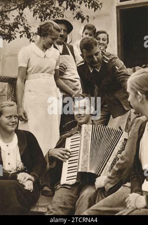 Nei quartieri, un soldato tedesco suona una fisarmonica, creando un'atmosfera vivace e piacevole. Questa scena cattura un momento di svago e di cameratismo tra le truppe, un netto contrasto con i rigori della vita militare. La musica e il divertimento condiviso sottolineano l'importanza del morale e dei momenti più leggeri che aiutano a sostenere i soldati in tempi difficili. Foto Stock