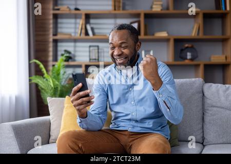 Vincitore online seduto a casa sul divano in salotto, felice uomo afro-americano di successo che celebra la vittoria, tenendo la mano in alto, ha ricevuto una notifica online al telefono, felice e soddisfatto. Foto Stock