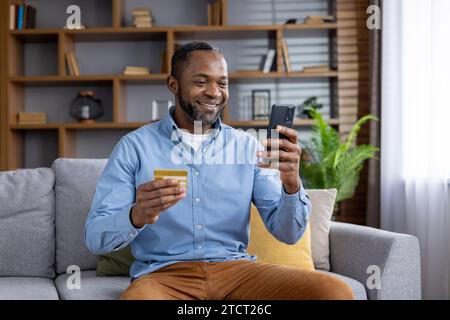 Uomo maturo gioioso a casa sul divano con telefono e carta di debito di credito in mano, uomo afro-americano soddisfatto in soggiorno sceglie regali nel negozio online, utilizza l'app sullo smartphone, trasferisce denaro. Foto Stock