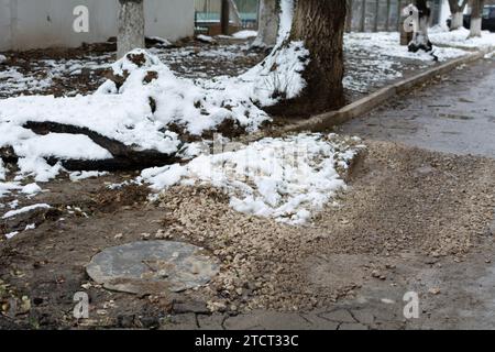 Lavori di riparazione su strada non completati. Portello fognario e asfalto rotto ricoperto di pietra frantumata. Sporcizia e neve su una strada rotta in una zona residenziale. Poco profondo Foto Stock