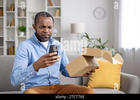 Insoddisfatto triste acquirente online, l'uomo ha ricevuto pacco danneggiato sbagliato, afroamericano seduto a casa sul divano in salotto con scatola di cartone nelle mani e telefono, scrivendo una recensione negativa. Foto Stock