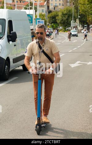 L'uomo (probabilmente locale, in uniforme da lavoro) viaggia / cavalca una strada elettrica con scooter elettrico / strada nr Palais des Festivals. Passeggiata sulla Croisette. Cannes Francia. (135) Foto Stock